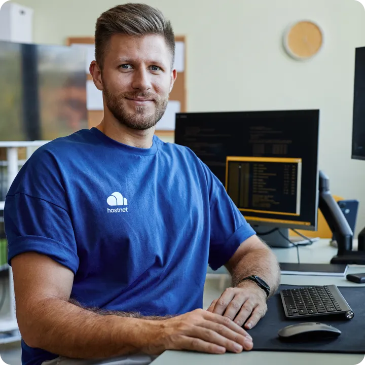 Man in blauw Hostnet-shirt aan een bureau met schermen vol code.